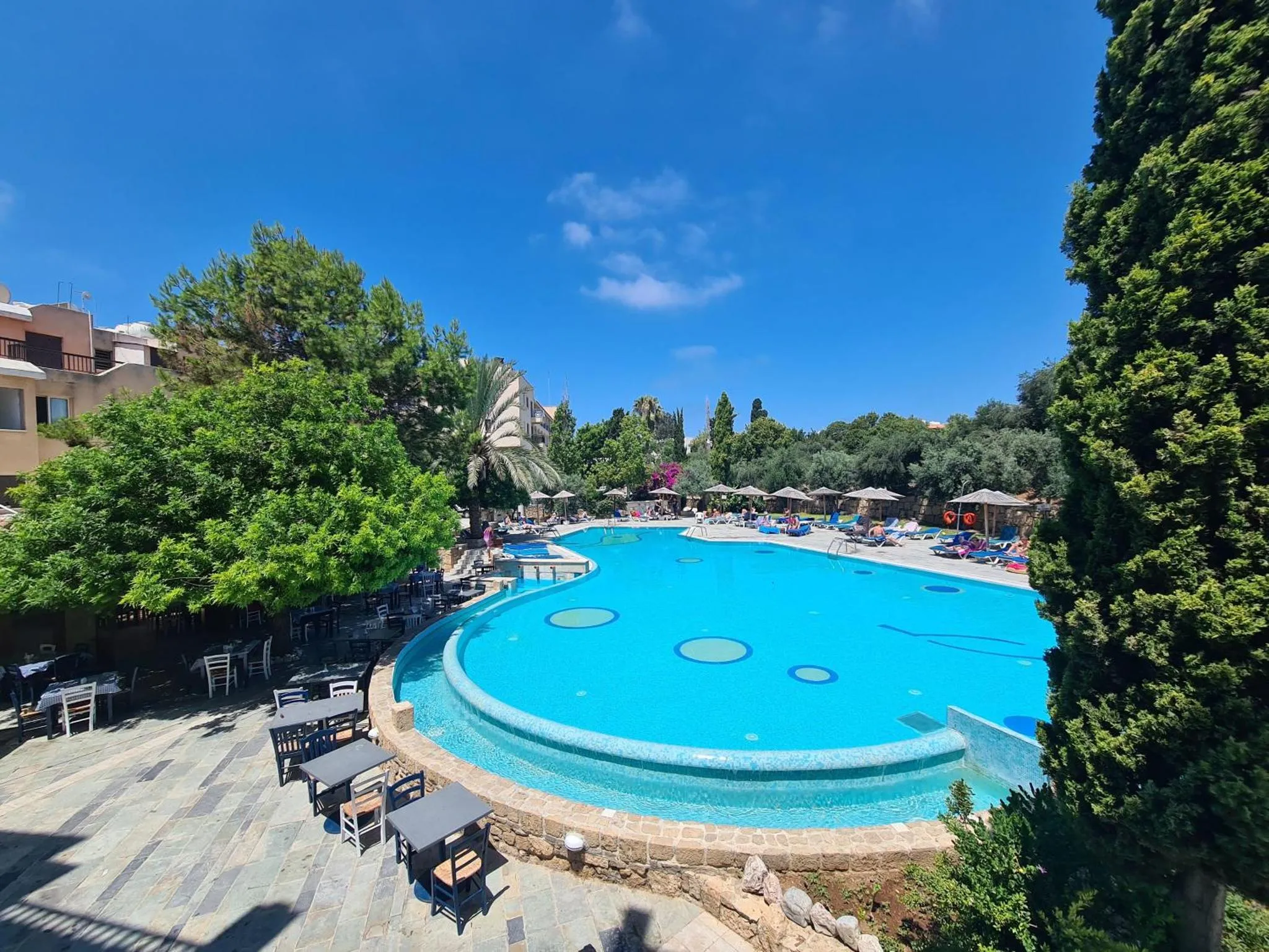 Une piscine turquoise entourée de transats et de parasols, avec des tables et des chaises sur une terrasse pavée.