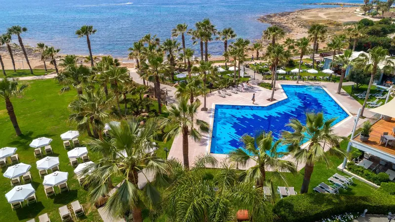 Vue aérienne d'une piscine bordée de palmiers et de transats sur une plage.