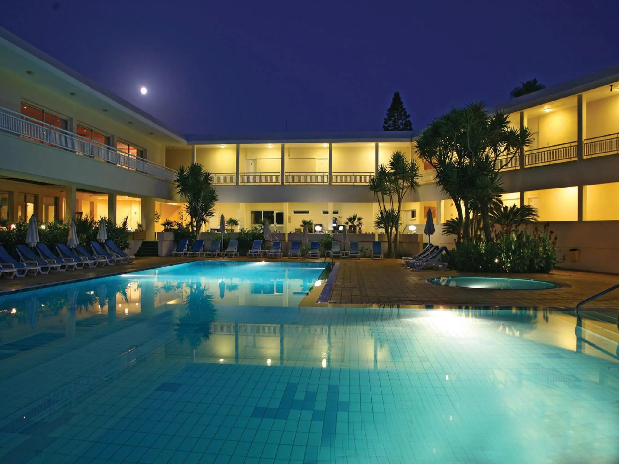Piscine illuminée au crépuscule avec transats et végétation tropicale.