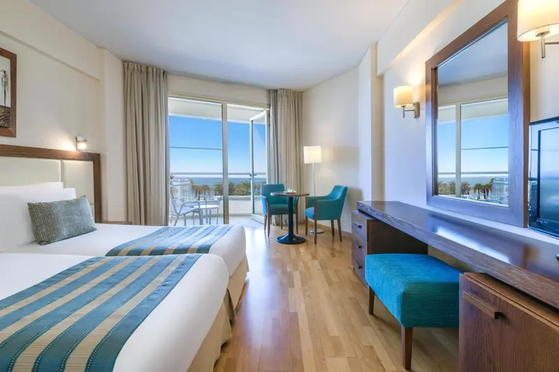 Chambre lumineuse avec lits jumeaux et vue sur la mer depuis un balcon.
