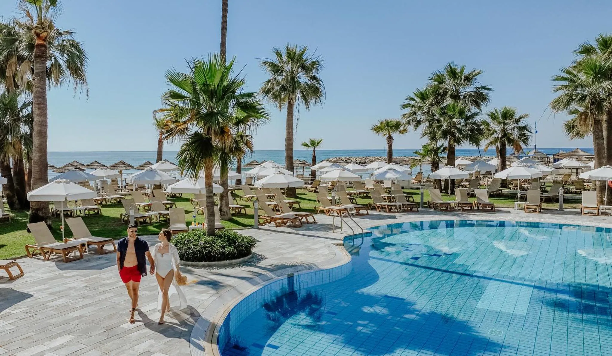 Une piscine bordée de transats et de palmiers, avec une plage en arrière-plan.