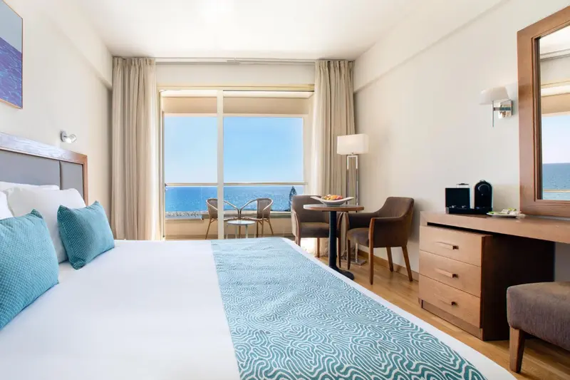 Chambre lumineuse avec un lit double et une vue sur la mer depuis un balcon.