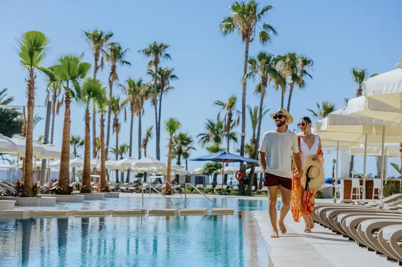 Un couple se promène près d'une piscine bordée de transats et de palmiers.
