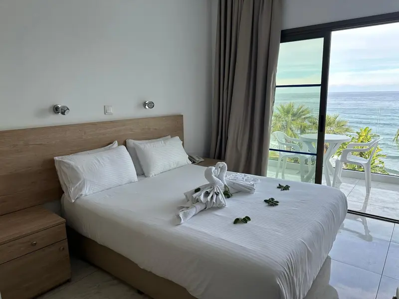 Chambre lumineuse avec un grand lit blanc et une vue sur la mer depuis le balcon.