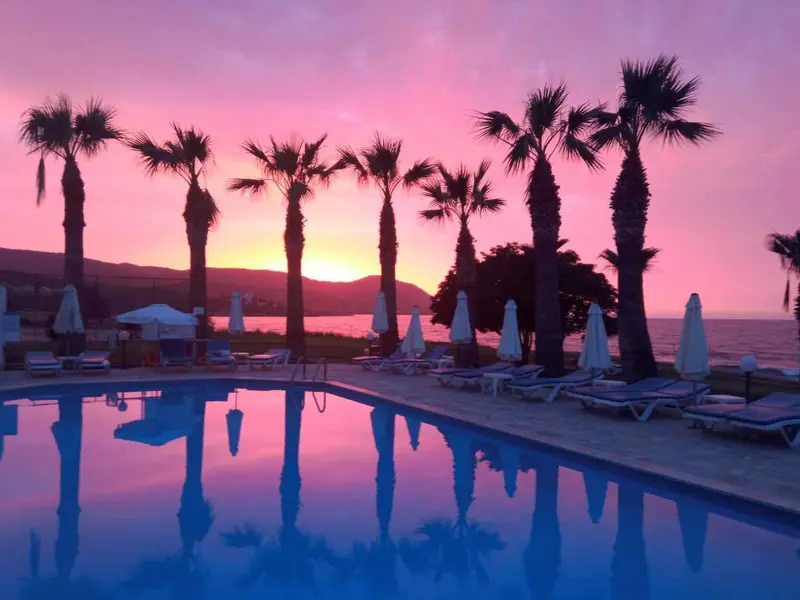 Une piscine reflète le ciel rose et orangé au coucher du soleil, bordée de transats et de palmiers.