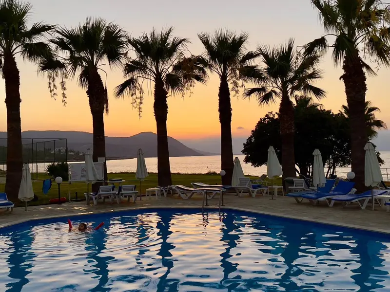 Transats bleus et parasols au bord d'une piscine avec des palmiers en arrière-plan au coucher du soleil.