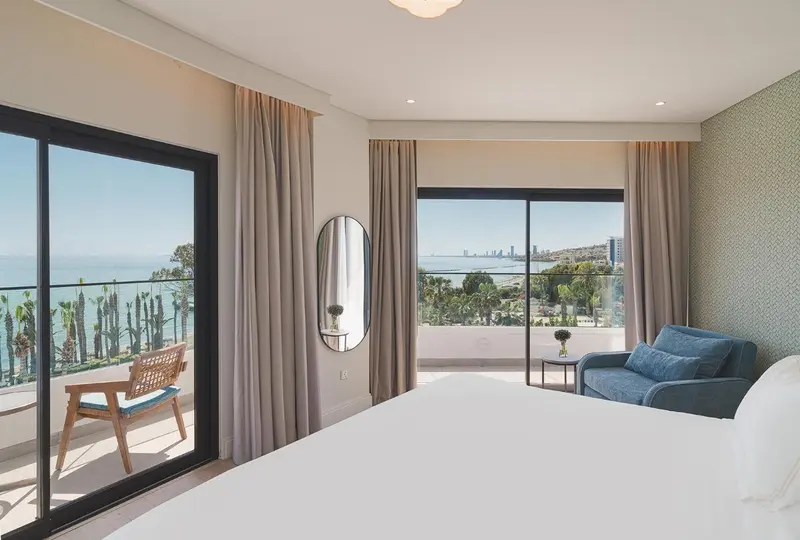 Chambre lumineuse avec un grand lit et un accès à un balcon offrant une vue sur la mer.
