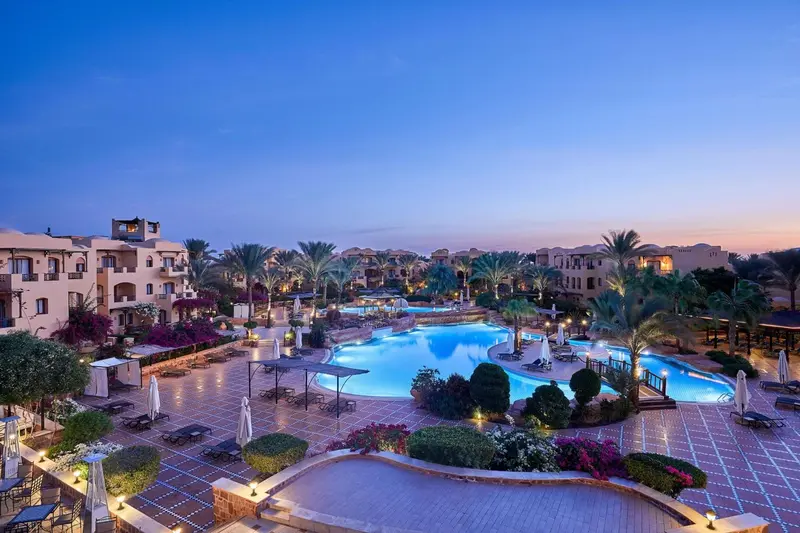 Vue d'un complexe hôtelier illuminé au crépuscule avec piscine et végétation tropicale.