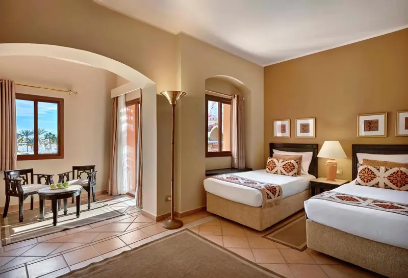 Chambre lumineuse avec deux lits et un coin salon près de la fenêtre.