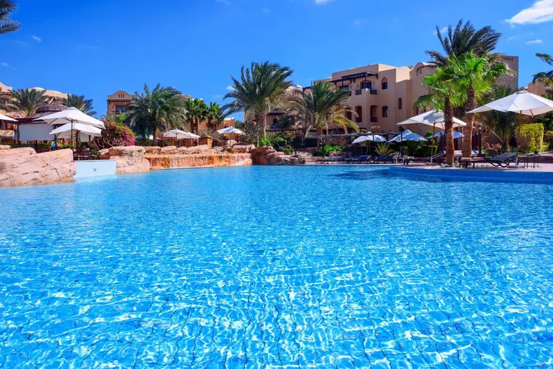 Une piscine turquoise bordée de transats et de palmiers sous un ciel bleu.