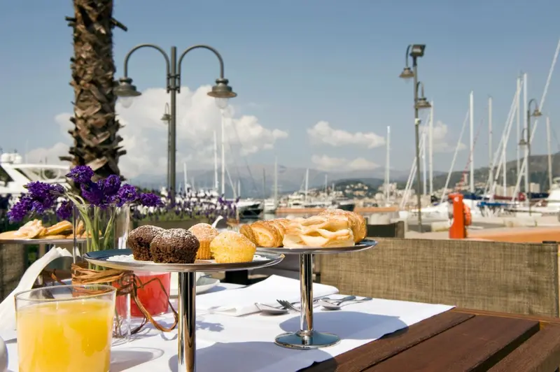 Une table est dressée avec des viennoiseries et une boisson fraîche, face à un port de plaisance ensoleillé.