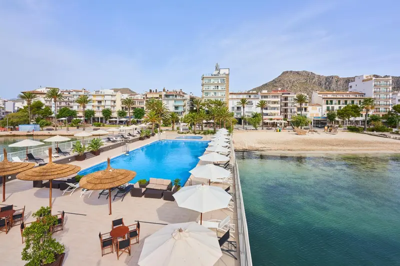 Vue aérienne d'une piscine d'hôtel bordée de transats et de parasols, avec la mer en arrière-plan et des bâtiments en hauteur.