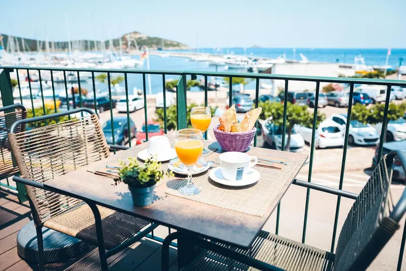 Une table est dressée pour le petit-déjeuner sur une terrasse ensoleillée avec vue sur un port de plaisance.