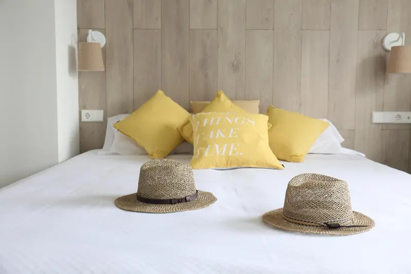 Un lit recouvert de draps blancs est orné de coussins jaunes et de deux chapeaux de paille.