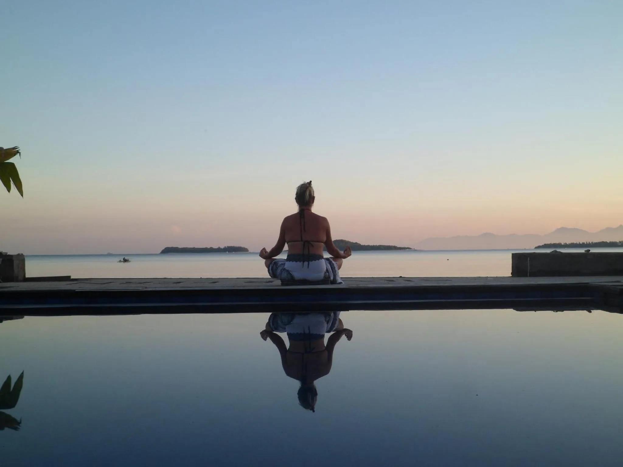 Une personne est assise en tailleur au bord d'une piscine face à la mer au coucher du soleil.