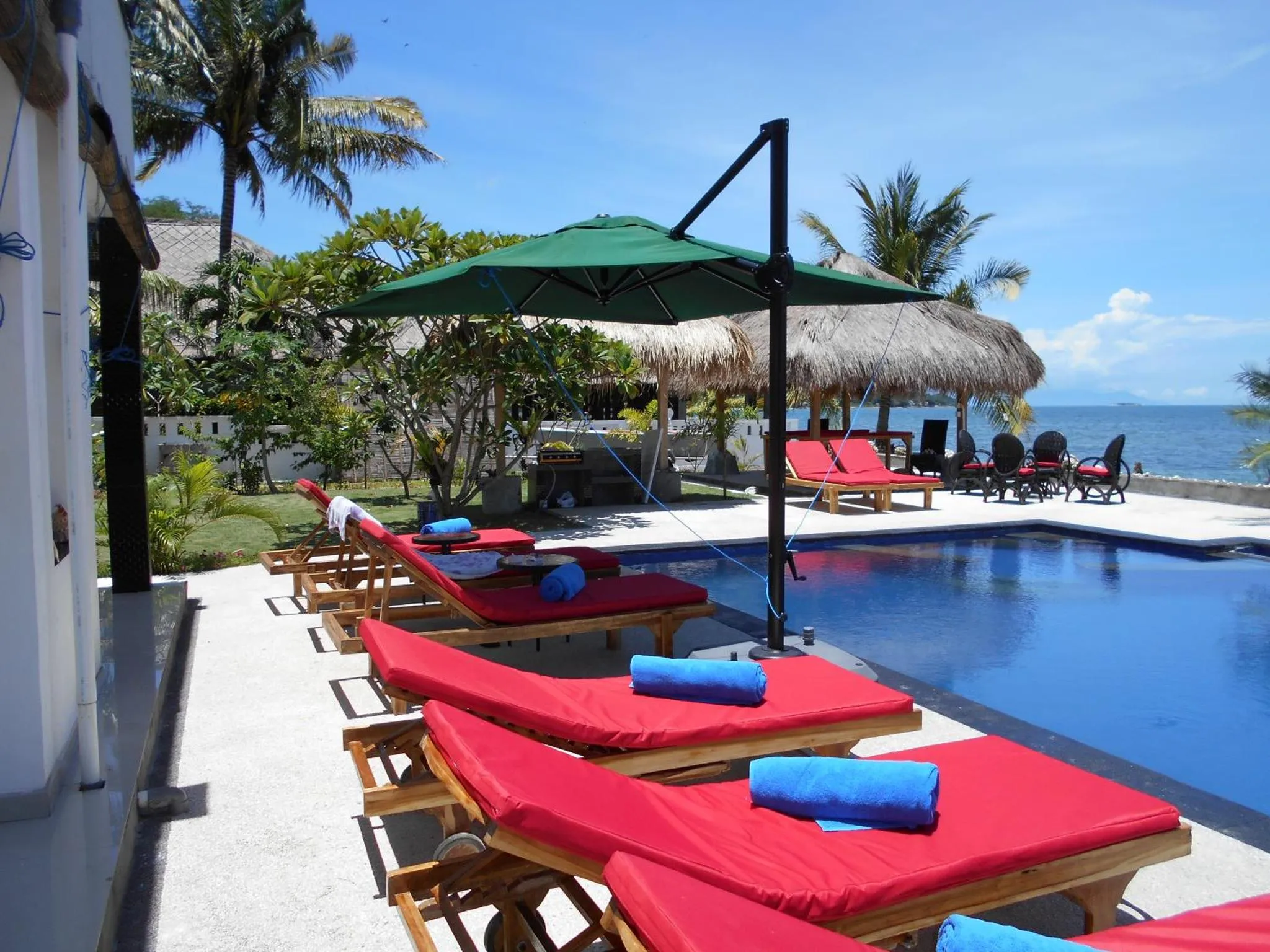 Transats rouges bordant une piscine turquoise sous des parasols verts, avec une vue sur la mer.