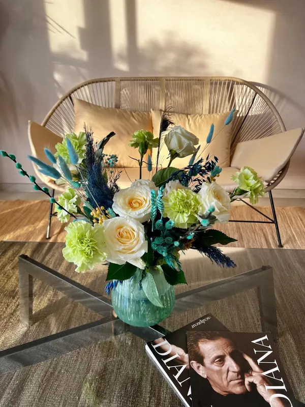 Composition florale colorée sur une table basse en verre devant un fauteuil en rotin.