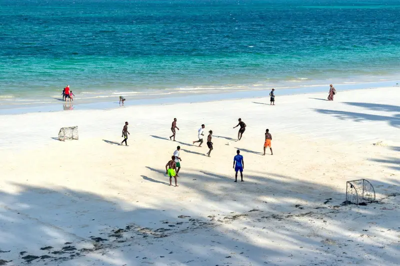 Des personnes jouent au football sur une plage de sable blanc bordée d'eau turquoise.