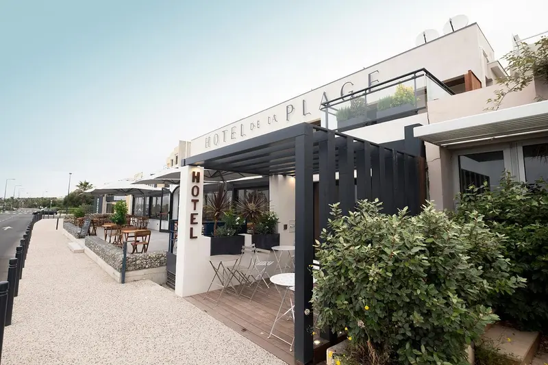 Façade d'un hôtel blanc avec une terrasse ombragée et des tables.