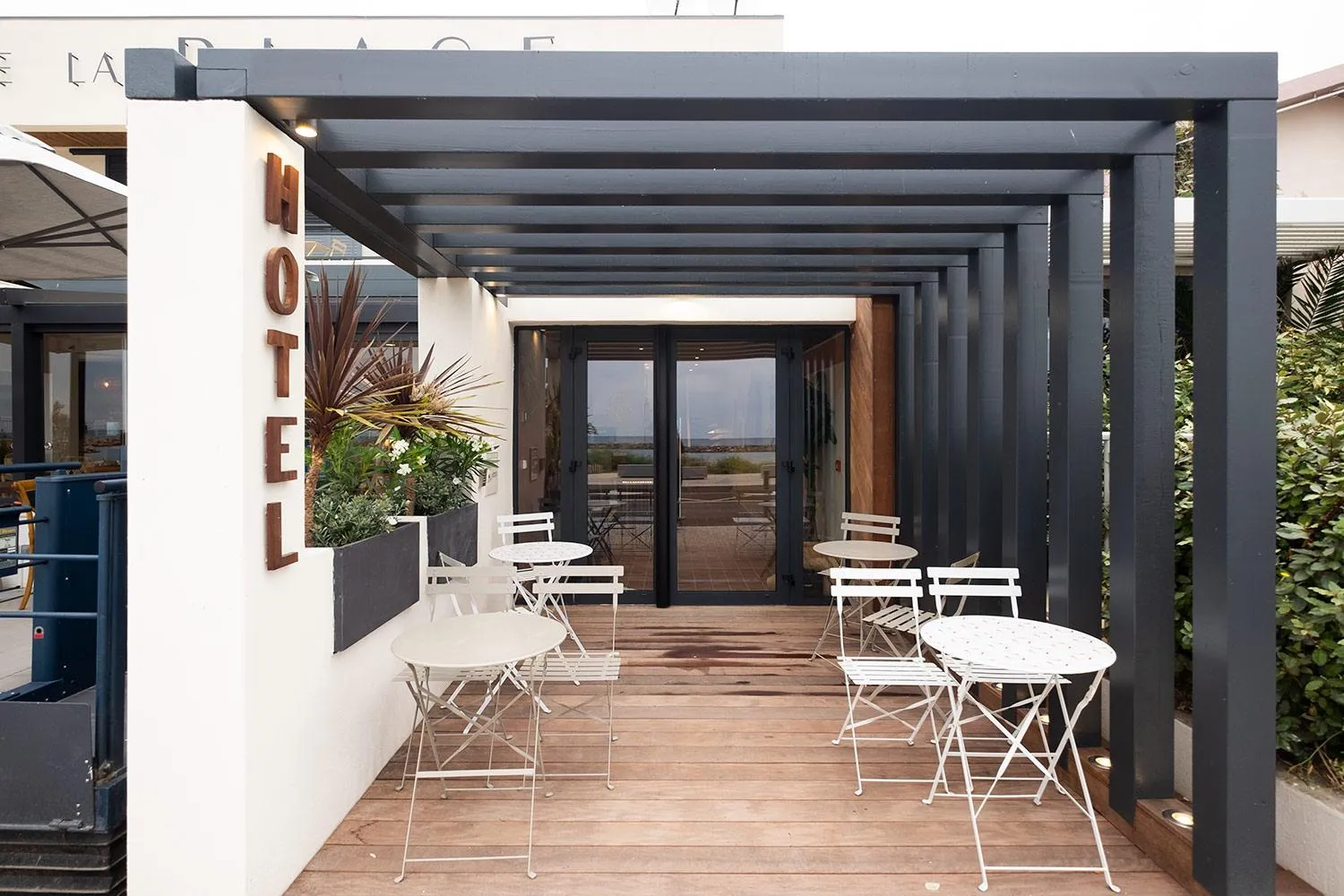 Terrasse ombragée avec tables et chaises blanches devant l'entrée d'un bâtiment.