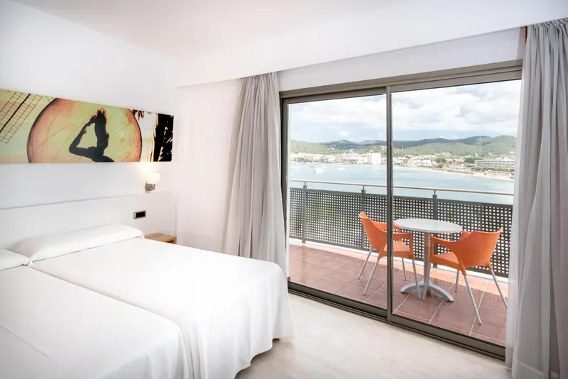 Chambre lumineuse avec un lit blanc et une vue sur la mer depuis un balcon.