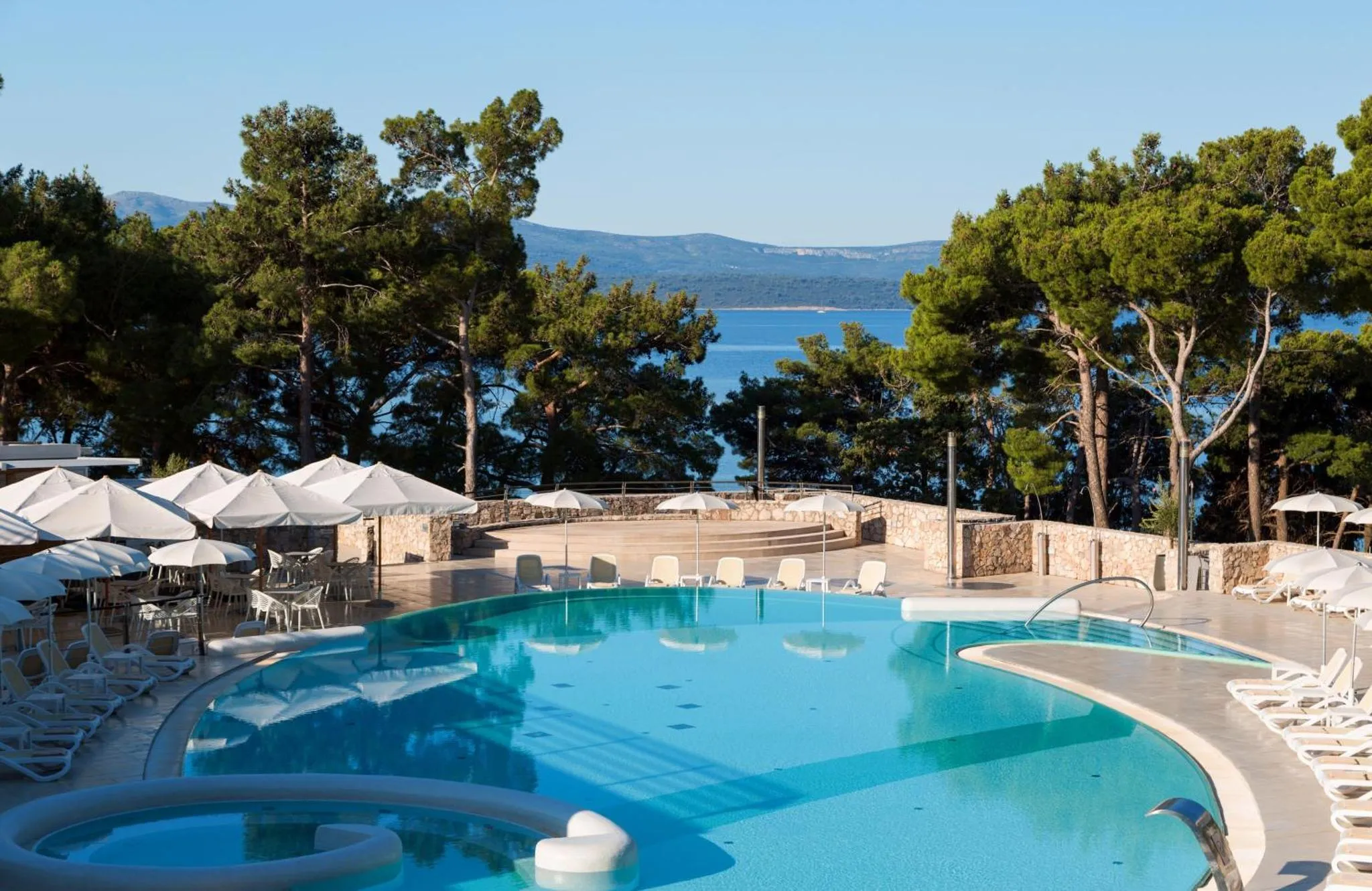 Une piscine bleue bordée de transats et de parasols offre une vue sur la mer et les collines verdoyantes.