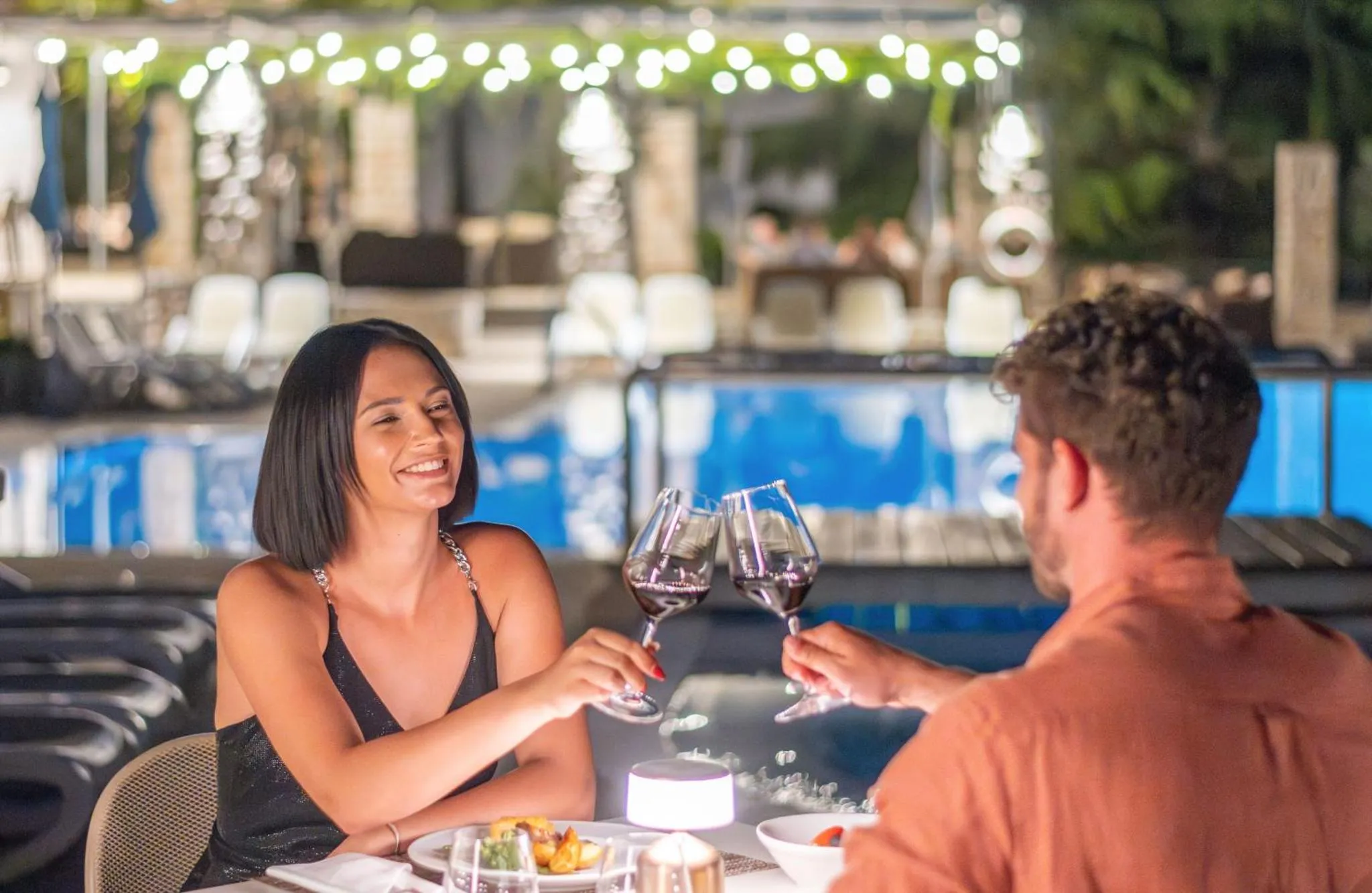 Un couple partage un moment de détente avec des verres de vin près d'une piscine illuminée.
