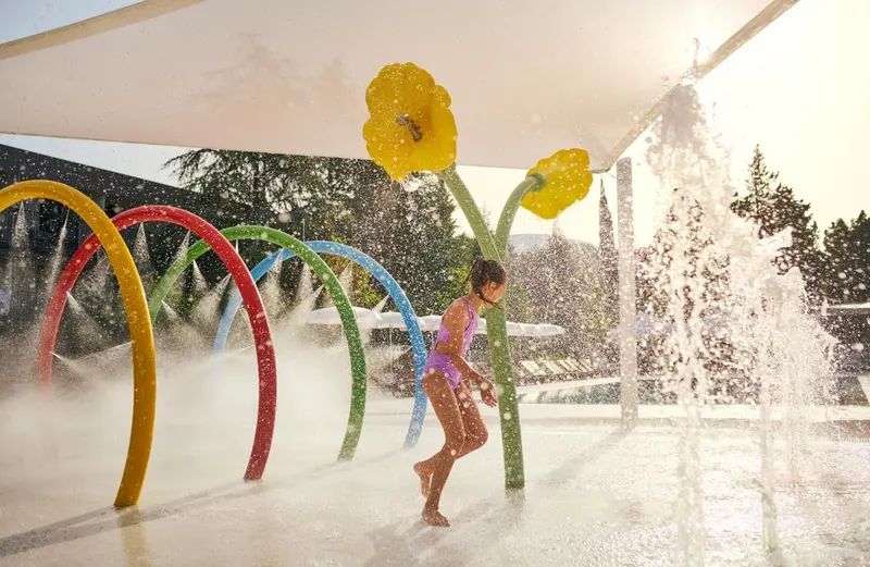 Une enfant joue dans un espace aquatique avec des jets d'eau et des structures colorées.