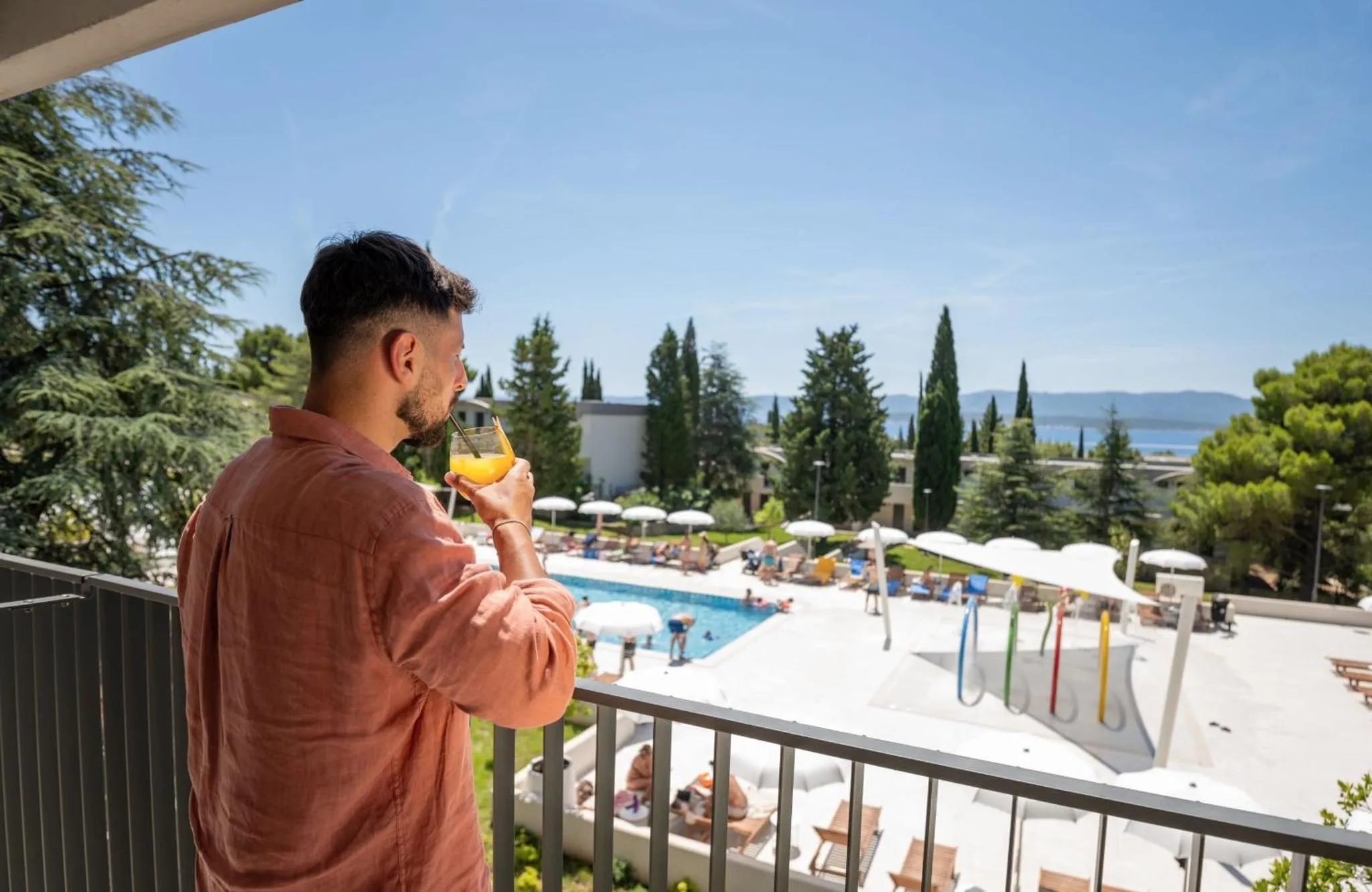 Un homme boit une boisson sur un balcon avec vue sur une piscine et la mer.