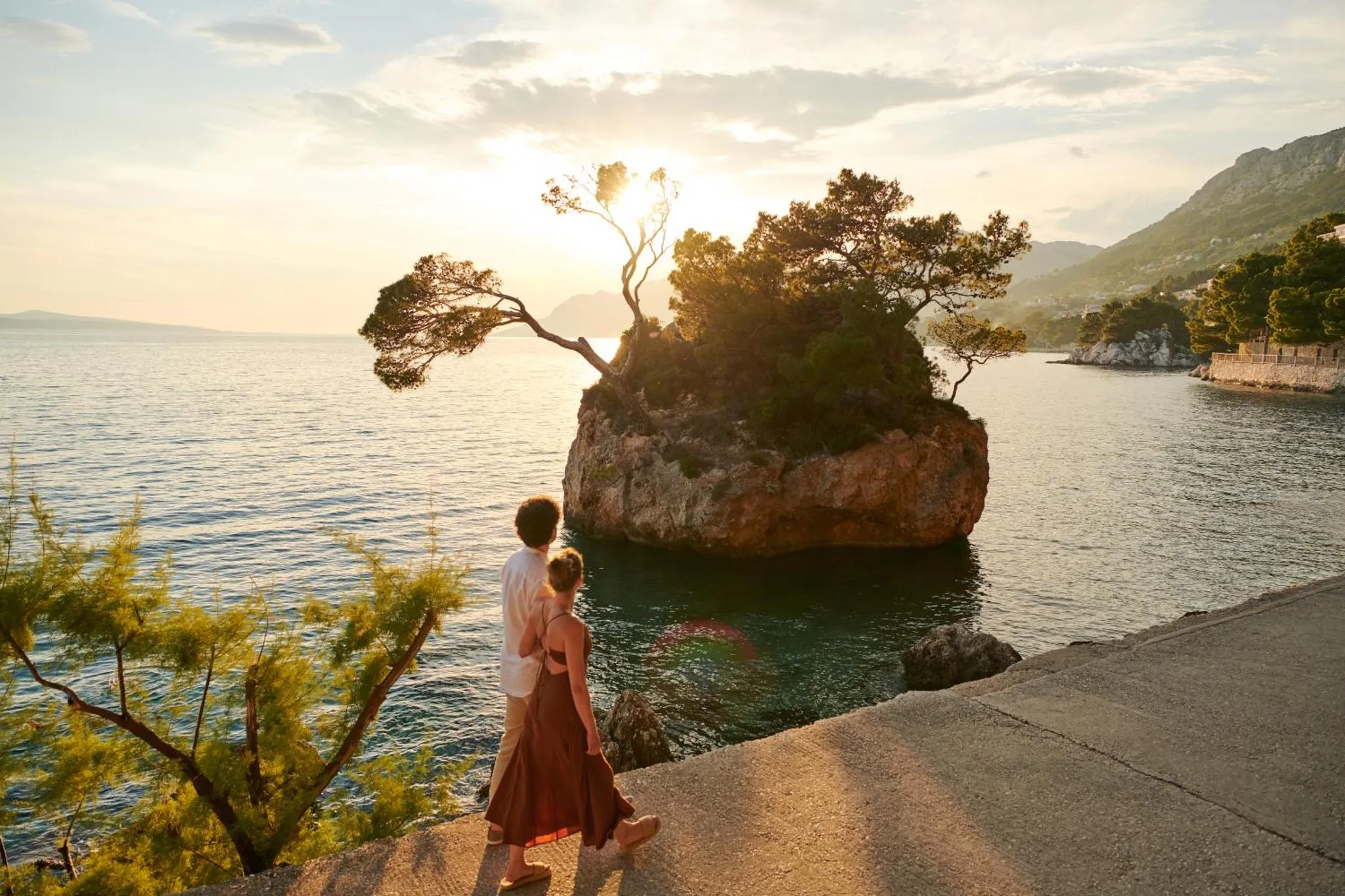 Un couple se tient sur une promenade rocheuse face à la mer au coucher du soleil.