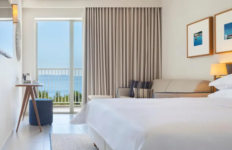 Chambre lumineuse avec un lit blanc et une vue sur la mer par de grandes baies vitrées.