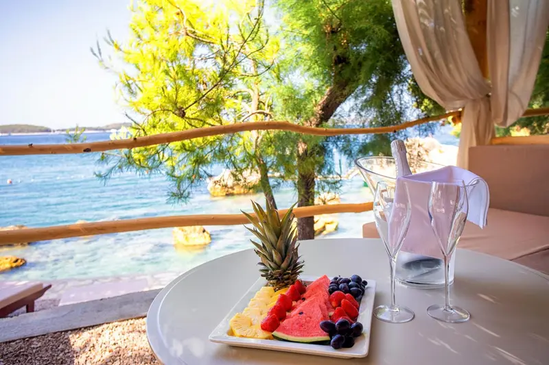 Une table ronde avec une assiette de fruits frais et des verres à vin sur une terrasse ensoleillée.
