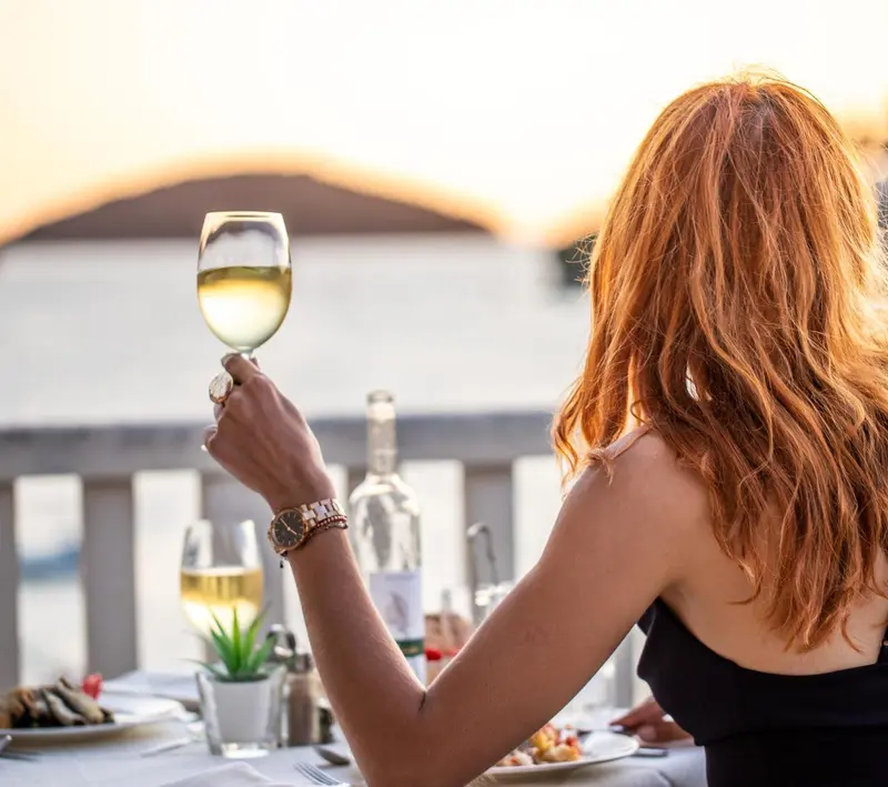 Une personne lève un verre de vin au coucher du soleil en terrasse.