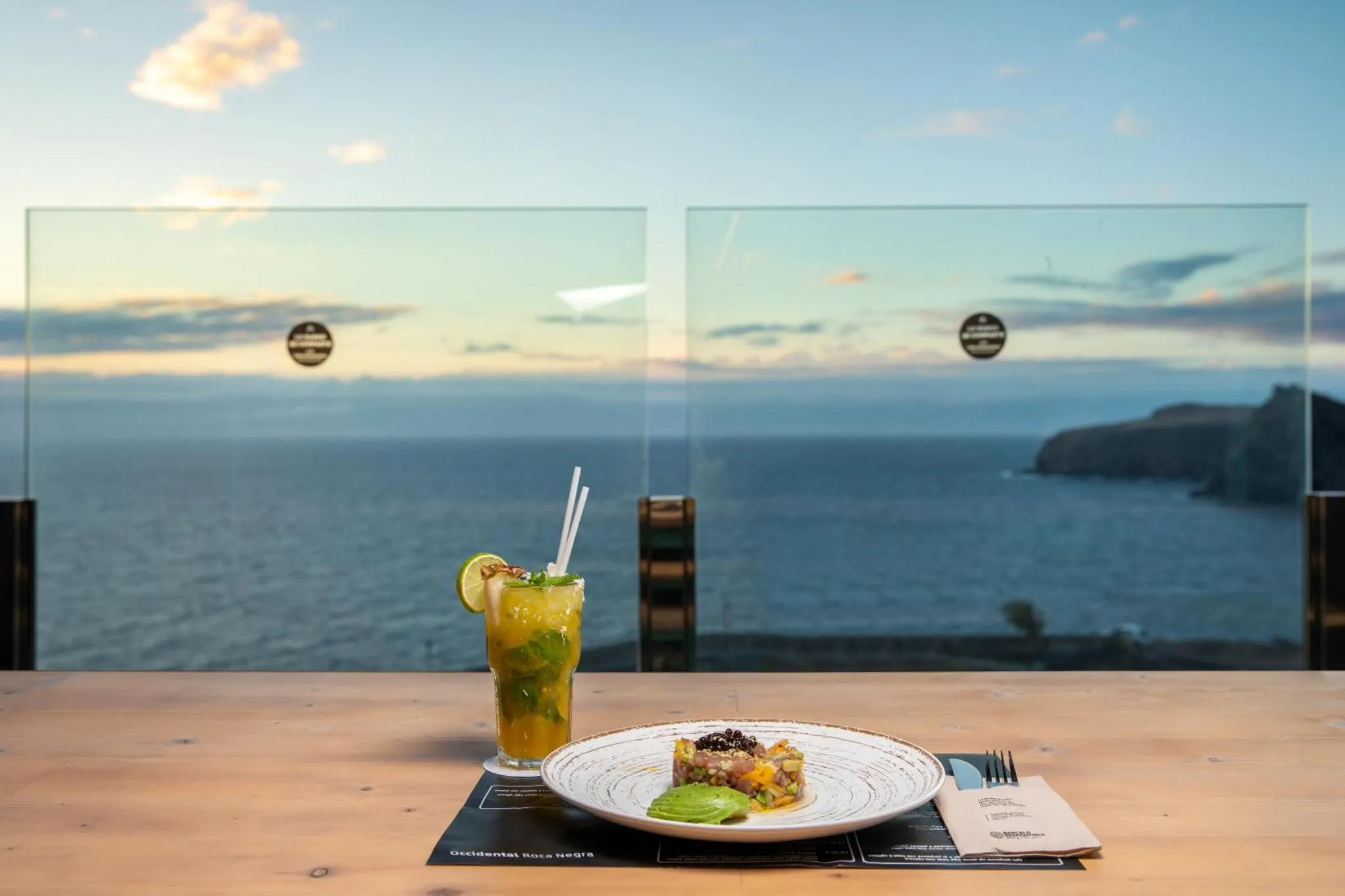 Une table en bois présente un plat et une boisson avec vue sur la mer.