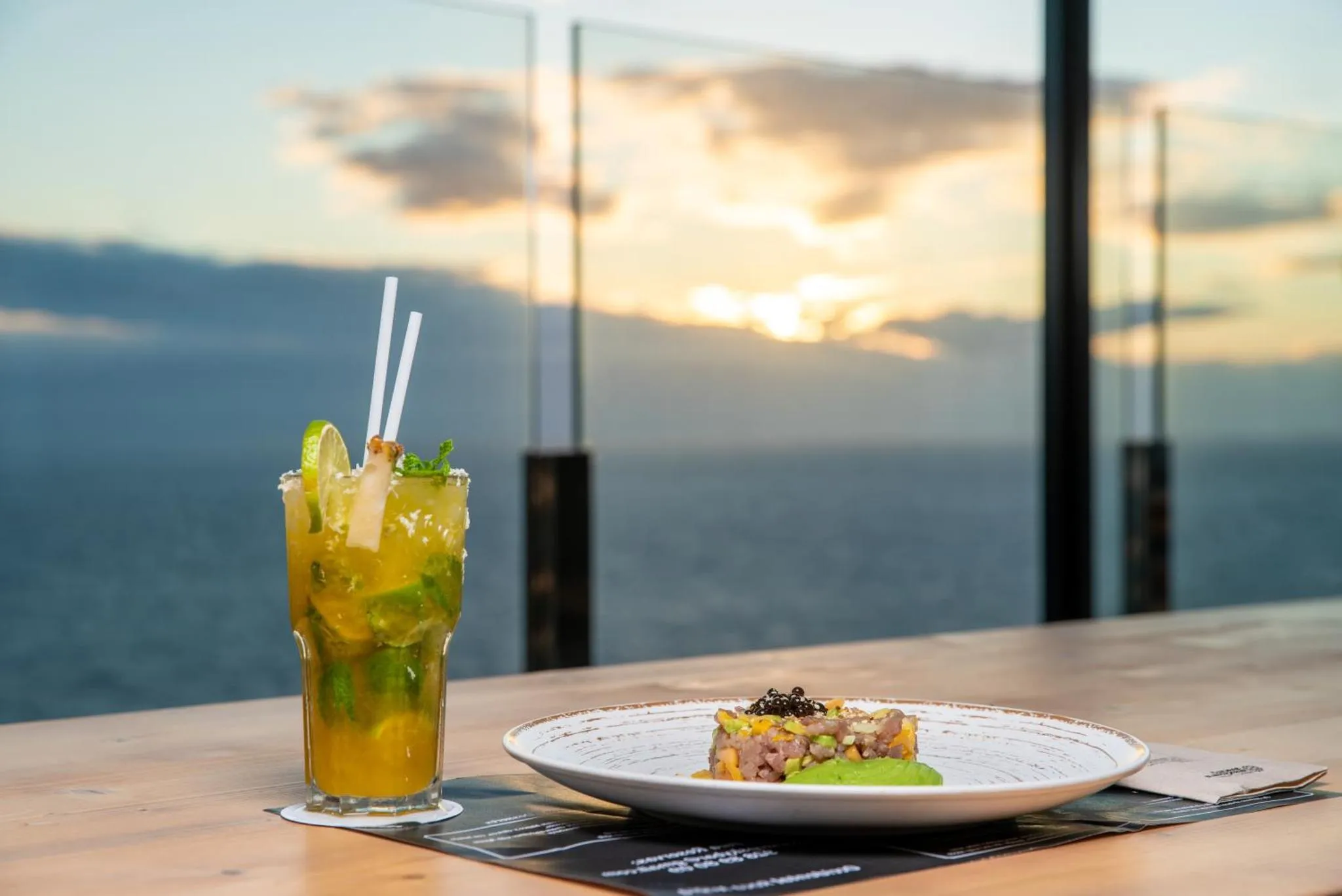Une table en bois présente des plats et une boisson rafraîchissante avec une vue sur la mer au coucher du soleil.