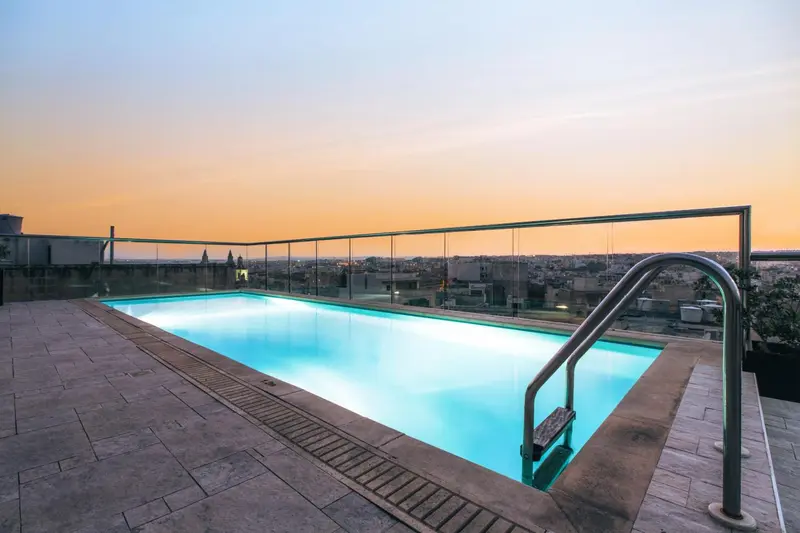 Une piscine rectangulaire sur un toit avec une vue sur la ville au coucher du soleil.