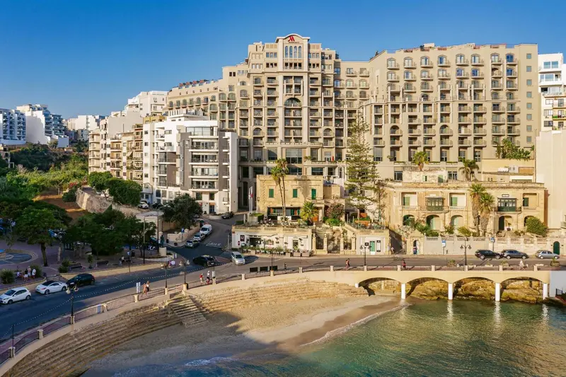 Vue d'un grand bâtiment blanc avec de nombreuses fenêtres et balcons, surplombant une promenade côtière et une petite plage.