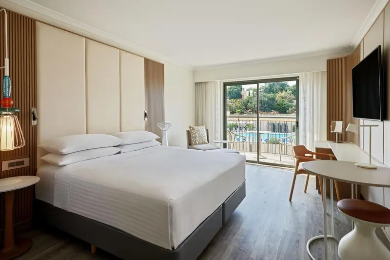 Chambre lumineuse avec un grand lit et une vue sur une piscine par une baie vitrée.
