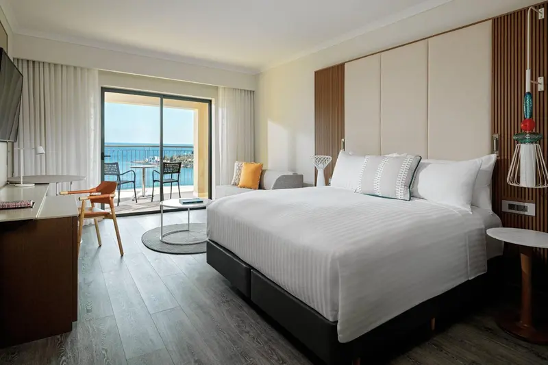 Chambre lumineuse avec un grand lit et un accès à un balcon donnant sur la mer.