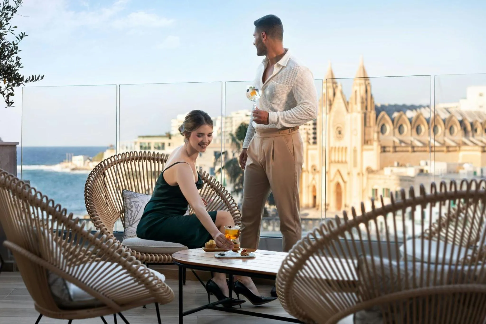 Un couple échangeant avec des cocktails sur une terrasse avec vue sur la mer et une ville.