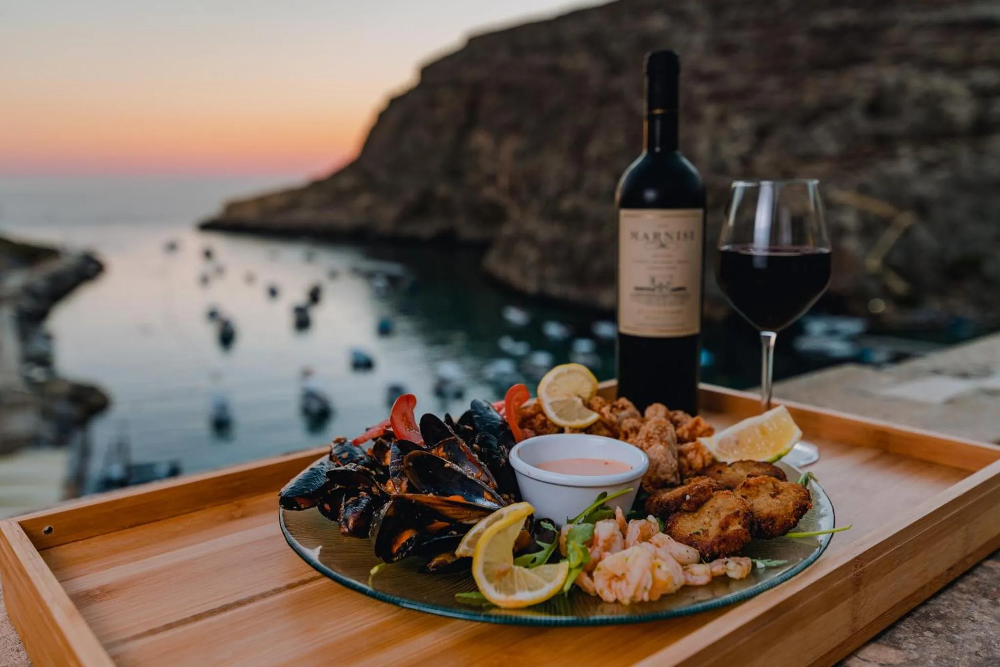 Une table en bois présente un plateau de fruits de mer et une bouteille de vin avec un verre, face à la mer au coucher du soleil.