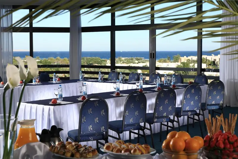 Une longue table est dressée avec des chaises dans une salle lumineuse donnant sur la mer.