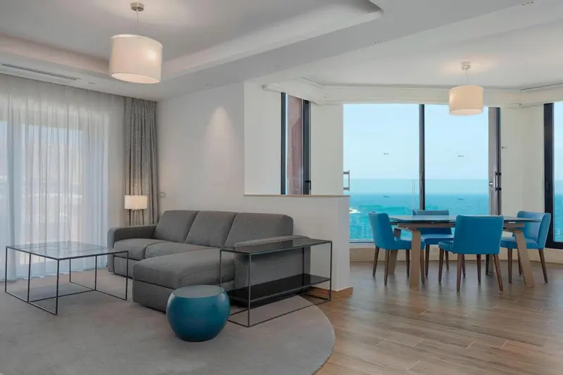 Un salon moderne avec canapé gris et table à manger près d'une grande fenêtre donnant sur la mer.