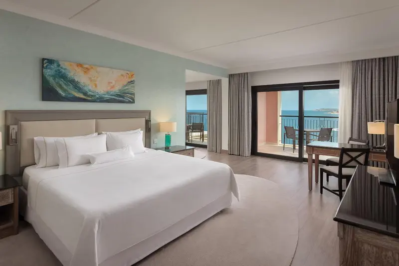Chambre lumineuse avec un grand lit et une vue sur la mer depuis un balcon.