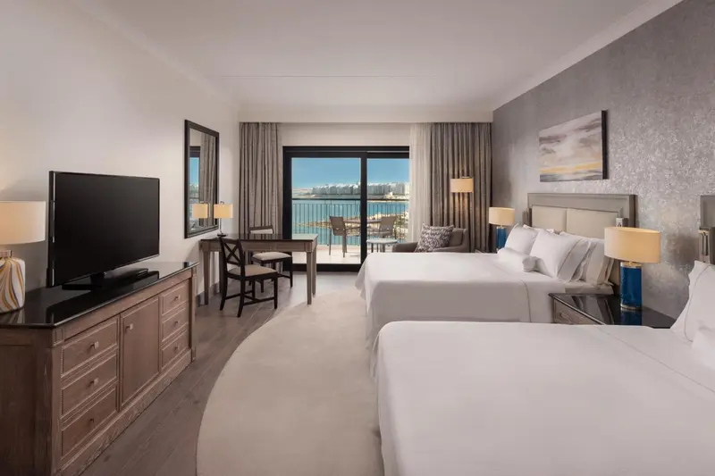 Chambre lumineuse avec deux lits et vue sur la mer par une baie vitrée.