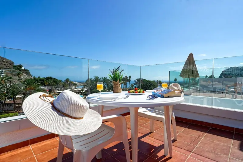 Une table ronde avec des boissons et des fruits sur une terrasse ensoleillée avec vue sur la mer.
