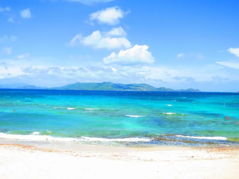 Vue d'une plage de sable blanc avec une mer turquoise et des îles à l'horizon sous un ciel bleu nuageux.