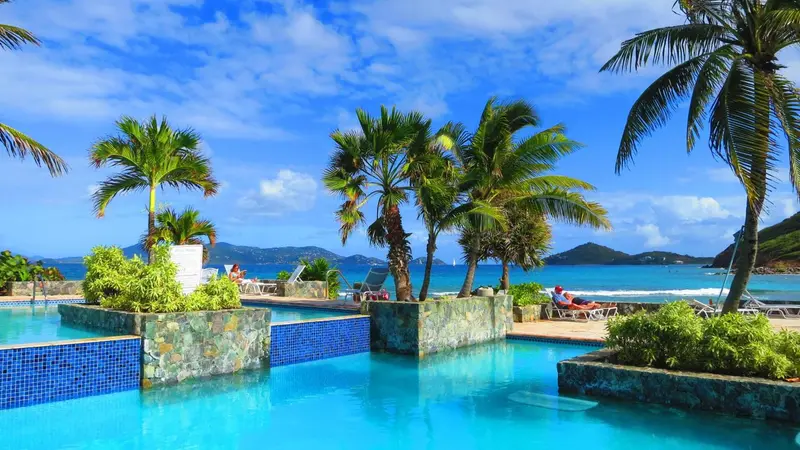 Une piscine turquoise bordée de végétation luxuriante offre une vue sur une plage de sable blanc et la mer.