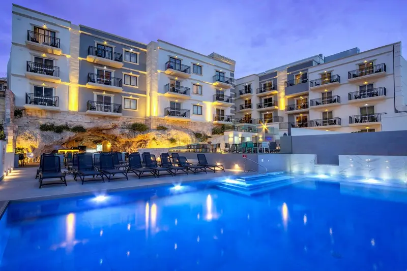 Bâtiment à plusieurs étages avec des balcons illuminés surplombant une piscine et des chaises longues.