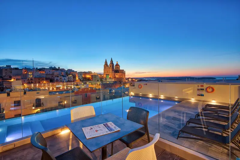 Une piscine à débordement avec des tables et des chaises offre une vue sur la ville illuminée au coucher du soleil.
